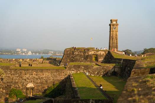 View of the historic Galle Fort Clock Tower and stone ramparts with people walking along the fortifications under a clear sky in Galle Southern Province Sri Lanka.