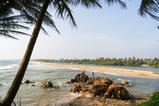 View of a tropical sandy beach and lagoon with people standing on large rocks under palm trees in Galle Southern Province Sri Lanka.