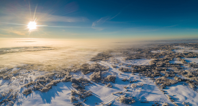Aerial view of a vast snow covered landscape in Latvia featuring frost covered trees and fields blanketed in white under a bright sun during the golden hour. Aerial view of a vast snow covered landscape in Latvia featuring frost covered trees and fields blanketed in white under a bright sun during the golden hour.