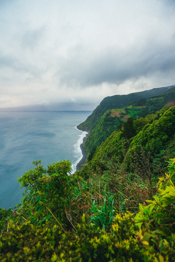 Aerial view of rugged cliffs cloaked in vibrant green vegetation meet the deep blue sea under a heavy overcast sky Azores Azores Portugal.