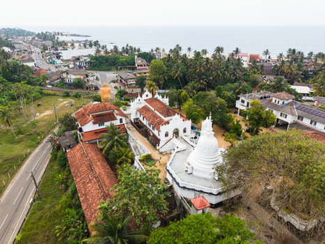 Aerial view of the Kumarakanda Kumara Maha Viharaya temple complex with a white stupa and traditional architecture surrounded by palm trees near the coast in Hikkaduwa Southern Province Sri Lanka. Aerial view of the Kumarakanda Kumara Maha Viharaya temple complex with a white stupa and traditional architecture surrounded by palm trees near the coast in Hikkaduwa Southern Province Sri Lanka.