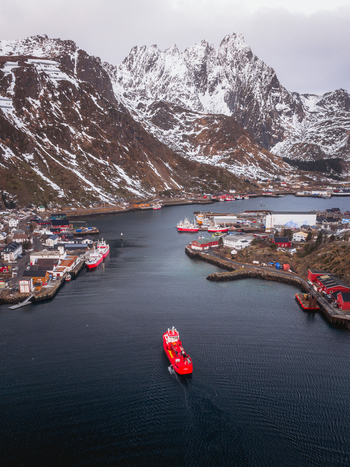 Aerial view of a vibrant red ship sailing through the calm dark waters towards the harbor nestled between snow dusted mountains Ballstad Nordland Norway. Aerial view of a vibrant red ship sailing through the calm dark waters towards the harbor nestled between snow dusted mountains Ballstad Nordland Norway.
