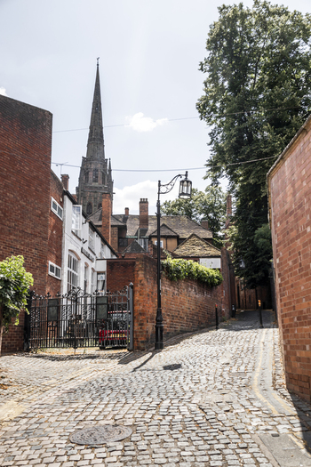 View of a historic cobblestone street leading towards a tall church spire with surrounding brick buildings and lush trees in Coventry England United Kingdom.