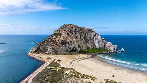 Aerial view of Morro Rock a massive volcanic plug standing on the sandy shoreline of the Pacific Ocean under a clear blue sky Morro Bay California United States.
