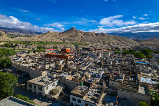Aerial view of a traditional village in the Himalayas with flat roofed stone houses a red monastery and colorful prayer flags set against arid mountain slopes in Nepal.