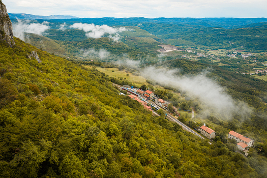 Aerial view of a railway station nestled in a lush green valley with low hanging clouds surrounding forests and winding roads in Buzet Istria County Croatia.