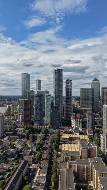 London United Kingdom 16 June 2026 Aerial view of Canary Wharf skyscrapers and modern office towers rising above residential neighborhoods under a cloudy blue sky.