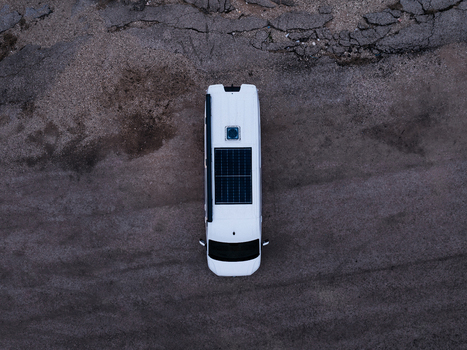 Aerial view of a white camper van with a solar panel on the roof parked on a gravel surface in Latvia.