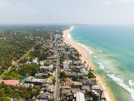 Aerial view of the coastal town with a long sandy beach turquoise ocean waves and buildings nestled among palm trees Hikkaduwa Southern Province Sri Lanka. Aerial view of the coastal town with a long sandy beach turquoise ocean waves and buildings nestled among palm trees Hikkaduwa Southern Province Sri Lanka.
