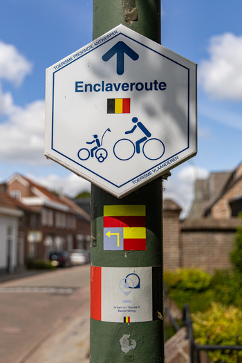 View of an Enclaveroute cycling path signpost featuring a hexagonal design with bicycle icons and the Belgian flag situated on a street in Baarle Hertog Flanders Belgium.