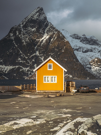 View of a vibrant yellow cabin nestled against a majestic snow dusted mountain range under a dramatic sky Hamn&oslash;y Nordland Norway.