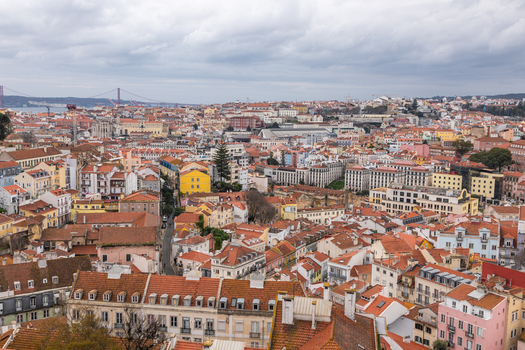 Aerial view of the historic cityscape of Lisbon featuring dense terracotta rooftops narrow streets and the 25 de Abril Bridge in the distance in Lisbon Lisbon Portugal.
