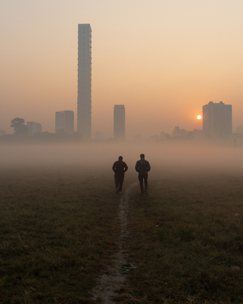 View of two people walking across a grassy field towards a city skyline during a foggy sunrise in Kolkata West Bengal India. View of two people walking across a grassy field towards a city skyline during a foggy sunrise in Kolkata West Bengal India.