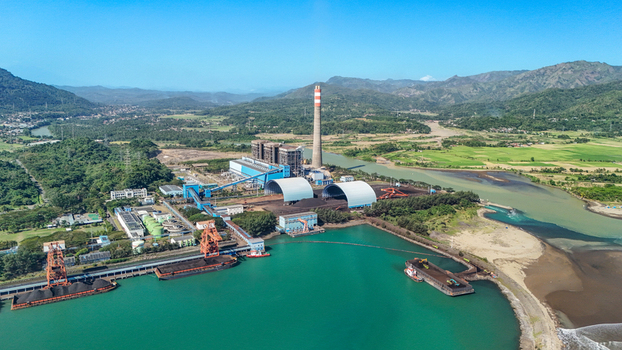 Aerial view of a coal fired power plant with industrial chimneys large storage domes and coal barges docked at the coastline in Indonesia.