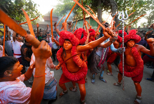Munshiganj Bangladesh 13 April 2025 View of vividly adorned devotees in orange paint and red garlands brandishing ceremonial swords and a trident during a street ritual. Munshiganj Bangladesh 13 April 2025 View of vividly adorned devotees in orange paint and red garlands brandishing ceremonial swords and a trident during a street ritual.