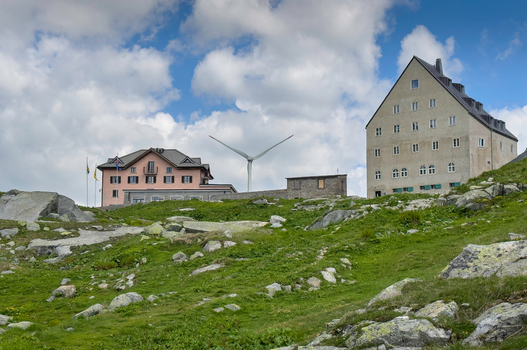 View of the Sankt Gotthard Pass buildings and a wind turbine on a rocky green hillside under a cloudy sky in Airolo Ticino Switzerland.