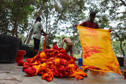Narayanganj Bangladesh 30 March 2026 View of vibrant yellow and red fabrics being manually processed creating a striking contrast against the muted tones of the surrounding outdoor workspace.