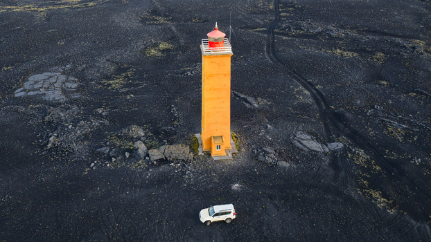 Aerial view of the yellow lighthouse standing in the middle of a vast black volcanic lava field with a white SUV parked nearby in Kirkjubaejarklaustur Skaftarhreppur Iceland. Aerial view of the yellow lighthouse standing in the middle of a vast black volcanic lava field with a white SUV parked nearby in Kirkjubaejarklaustur Skaftarhreppur Iceland.
