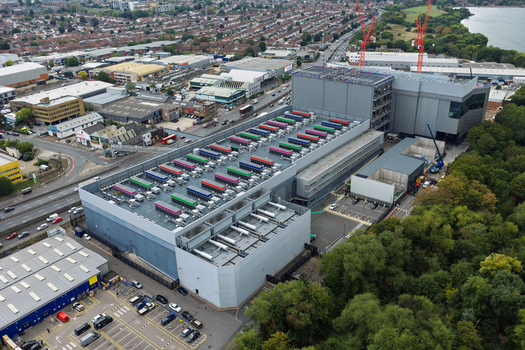 Aerial view of a large data center facility featuring colorful cooling units on the roof situated next to a busy road and a wooded area in the United Kingdom.