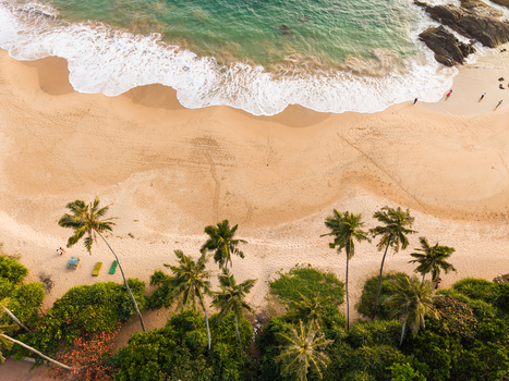 Aerial view of a sandy beach with turquoise waves crashing on the shore palm trees and people walking along the coastline in Galle Southern Province Sri Lanka.