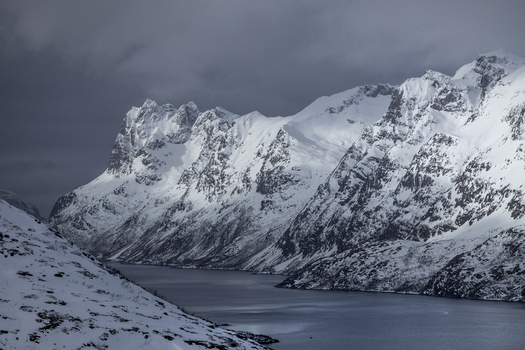 View of snow capped mountain peaks rising steeply above the dark waters of a fjord under a dramatic overcast sky Ersfjord Troms Norway. View of snow capped mountain peaks rising steeply above the dark waters of a fjord under a dramatic overcast sky Ersfjord Troms Norway.