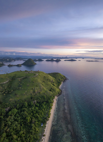 Aerial view of Labuan Bajo coastline featuring lush green hills a pristine sandy beach and calm turquoise waters under a soft sunset sky in Komodo East Nusa Tenggara Indonesia. Aerial view of Labuan Bajo coastline featuring lush green hills a pristine sandy beach and calm turquoise waters under a soft sunset sky in Komodo East Nusa Tenggara Indonesia.