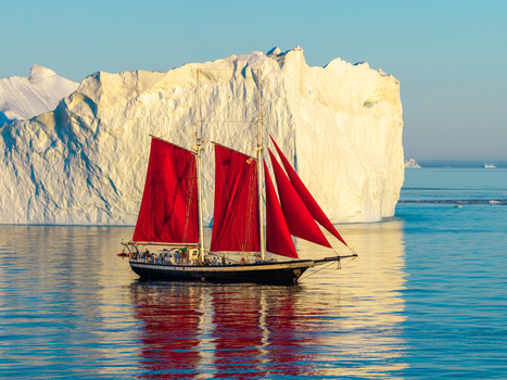 Aerial view of a sailboat with striking red sails navigating calm waters in front of a massive white iceberg in Greenland. Aerial view of a sailboat with striking red sails navigating calm waters in front of a massive white iceberg in Greenland.