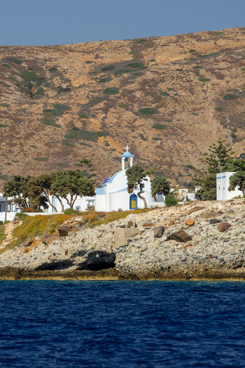 View of a traditional white church with a blue dome and a Greek flag situated on a rocky coastline with sparse trees and arid hills in Fira Greece. View of a traditional white church with a blue dome and a Greek flag situated on a rocky coastline with sparse trees and arid hills in Fira Greece.