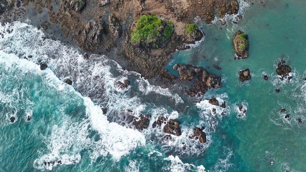 Aerial view of the rugged coastline with dark volcanic rocks crashing turquoise waves and lush green vegetation along the shore in Indonesia. Aerial view of the rugged coastline with dark volcanic rocks crashing turquoise waves and lush green vegetation along the shore in Indonesia.