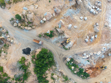 Aerial view of a mining site with an excavator and a dump truck operating on rocky terrain amidst dirt paths and sparse vegetation in Bukit Tambun Penang Malaysia. Aerial view of a mining site with an excavator and a dump truck operating on rocky terrain amidst dirt paths and sparse vegetation in Bukit Tambun Penang Malaysia.