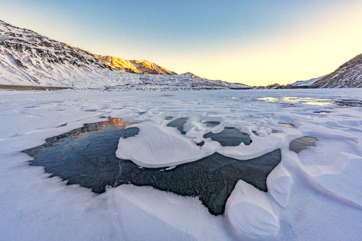 View of a frozen lake with patches of ice and snow surrounded by snow covered mountains under a clear sunset sky in Madesimo Lombardy Italy. View of a frozen lake with patches of ice and snow surrounded by snow covered mountains under a clear sunset sky in Madesimo Lombardy Italy.