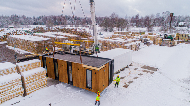 Aerial view of a modular house being lifted by a crane in a snowy lumber yard with stacks of timber and workers in high visibility jackets in Latvia. Aerial view of a modular house being lifted by a crane in a snowy lumber yard with stacks of timber and workers in high visibility jackets in Latvia.