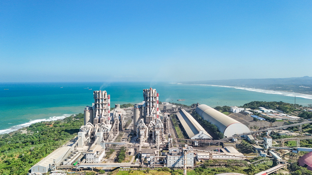 Aerial view of a large industrial cement plant featuring tall processing silos and storage warehouses situated along the coastline with turquoise ocean waters and a clear blue sky in Indonesia.