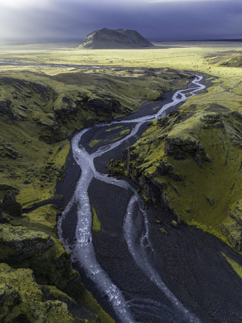 Aerial view of Petursey mountain and a braided glacial river winding through a mossy green canyon under a dramatic cloudy sky Skeidflotur Myrdalshreppur Iceland.
