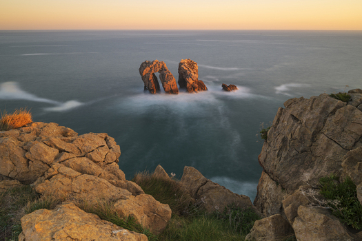 View of the Urros de Liencres rock formations and natural arch with long exposure water and golden sunset light from the cliffs Liencres Cantabria Spain. View of the Urros de Liencres rock formations and natural arch with long exposure water and golden sunset light from the cliffs Liencres Cantabria Spain.
