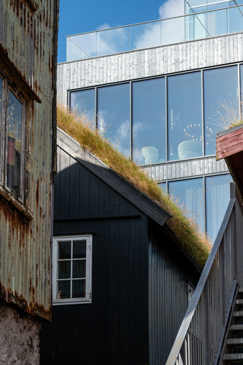 View of a traditional black wooden house with a grass roof standing next to a modern glass building in the Faroe Islands. View of a traditional black wooden house with a grass roof standing next to a modern glass building in the Faroe Islands.