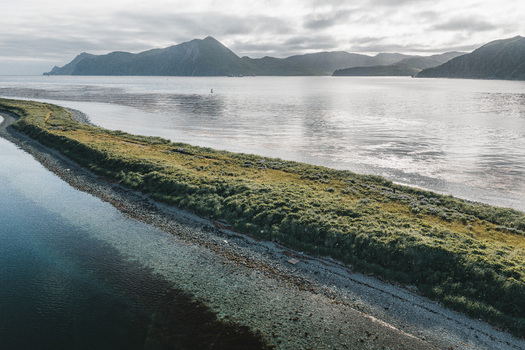 Aerial view of the narrow grassy spit at Captains Bay with calm waters and rugged mountains under an overcast sky in Unalaska Alaska United States. Aerial view of the narrow grassy spit at Captains Bay with calm waters and rugged mountains under an overcast sky in Unalaska Alaska United States.