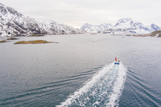 Aerial view of a boat cruising through the calm waters of a fjord in Lofoten surrounded by snow capped mountains under a bright sky Nordland Norway.