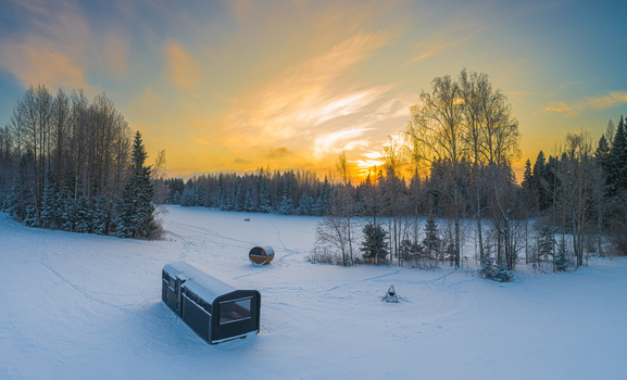 Aerial view of a small modern cabin and a barrel sauna in a snowy forest landscape during a golden sunset in Latvia. Aerial view of a small modern cabin and a barrel sauna in a snowy forest landscape during a golden sunset in Latvia.