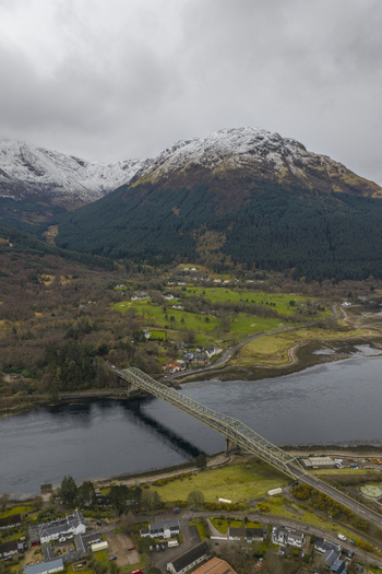 Aerial view of Ballachulish Bridge crossing the water with snow capped mountains and green fields in Ballachulish Scotland United Kingdom.