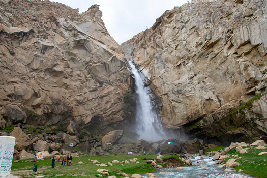 View of Khamosh Waterfall cascading down steep rocky cliffs into a stream with people on a grassy field Skardu Gilgit Pakistan.