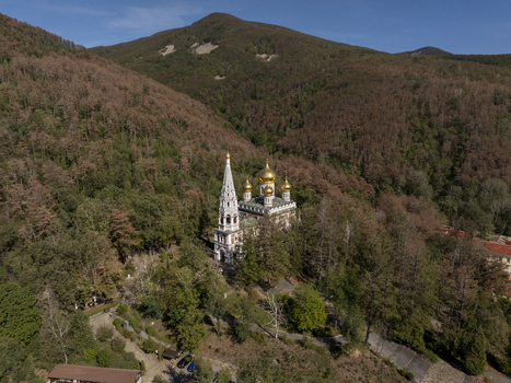 Aerial view of Shipka Memorial Church nestled in a dense forest with golden domes and a tall bell tower against a mountain backdrop in Shipka Stara Zagora Bulgaria. Aerial view of Shipka Memorial Church nestled in a dense forest with golden domes and a tall bell tower against a mountain backdrop in Shipka Stara Zagora Bulgaria.