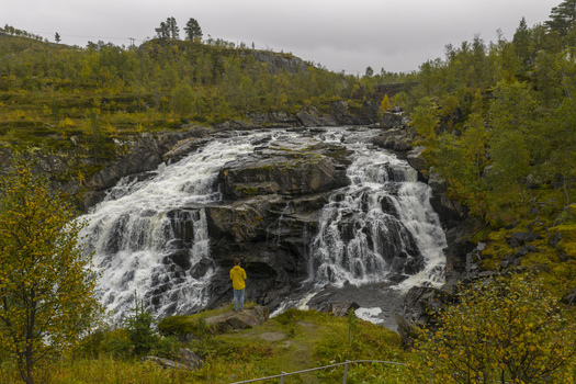 Aerial view of Voringfossen waterfall with a person in a yellow jacket standing on a rock surrounded by autumn foliage in Eidfjord Municipality Vestland Norway. Aerial view of Voringfossen waterfall with a person in a yellow jacket standing on a rock surrounded by autumn foliage in Eidfjord Municipality Vestland Norway.