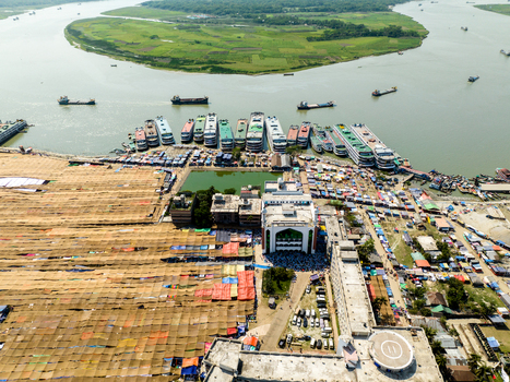 Aerial view of a bustling riverfront with ferries docked alongside a vibrant trading area contrasting with the calm river and distant green island Charmonai Barisal Division Bangladesh. Aerial view of a bustling riverfront with ferries docked alongside a vibrant trading area contrasting with the calm river and distant green island Charmonai Barisal Division Bangladesh.