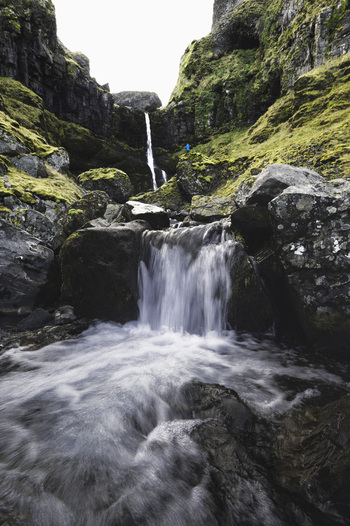 View of Bugsfoss waterfall cascading over dark volcanic rocks into a rushing stream with mossy cliffs and a person in a blue jacket Olafsvik Snaefellsbaer Iceland.