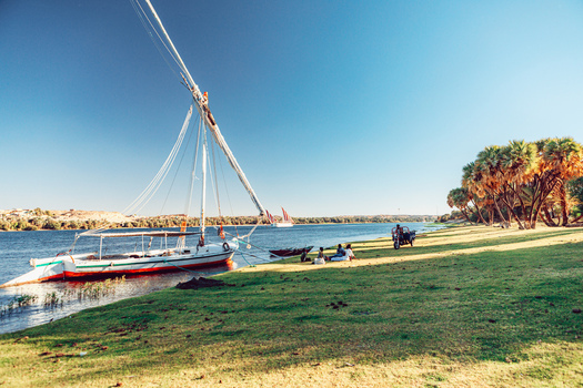 View of a traditional felucca sailboat moored on the grassy banks of the Nile river with people resting under palm trees in Aswan Aswan Governorate Egypt. View of a traditional felucca sailboat moored on the grassy banks of the Nile river with people resting under palm trees in Aswan Aswan Governorate Egypt.