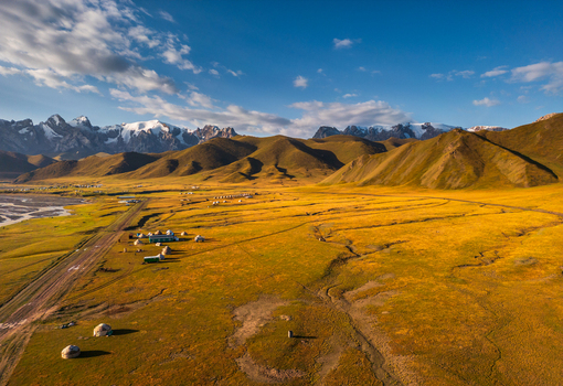 Aerial view of Kol Suu featuring traditional yurts on the golden mountain landscape with snow capped peaks under a blue sky in At Bashi District Naryn Region Kyrgyzstan. Aerial view of Kol Suu featuring traditional yurts on the golden mountain landscape with snow capped peaks under a blue sky in At Bashi District Naryn Region Kyrgyzstan.