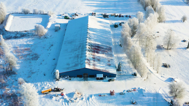 Aerial view of a blue agricultural building surrounded by snow covered fields and frost covered trees in Latvija. Aerial view of a blue agricultural building surrounded by snow covered fields and frost covered trees in Latvija.