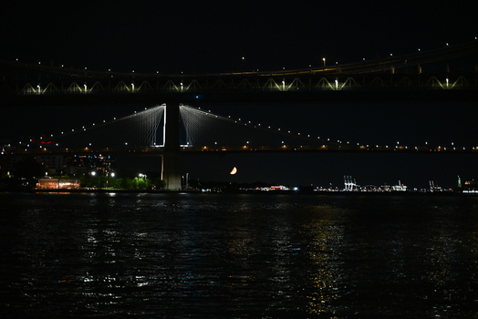 View of the Brooklyn Bridge illuminated at night over the East River with city lights reflecting on the water and a crescent moon New York New York United States. View of the Brooklyn Bridge illuminated at night over the East River with city lights reflecting on the water and a crescent moon New York New York United States.