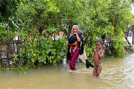 Banshkhali Upazila Chittagong Division Bangladesh 10 July 2026 View of a woman carrying a child and a young boy wading through floodwaters in a rural area surrounded by lush green vegetation. Banshkhali Upazila Chittagong Division Bangladesh 10 July 2026 View of a woman carrying a child and a young boy wading through floodwaters in a rural area surrounded by lush green vegetation.